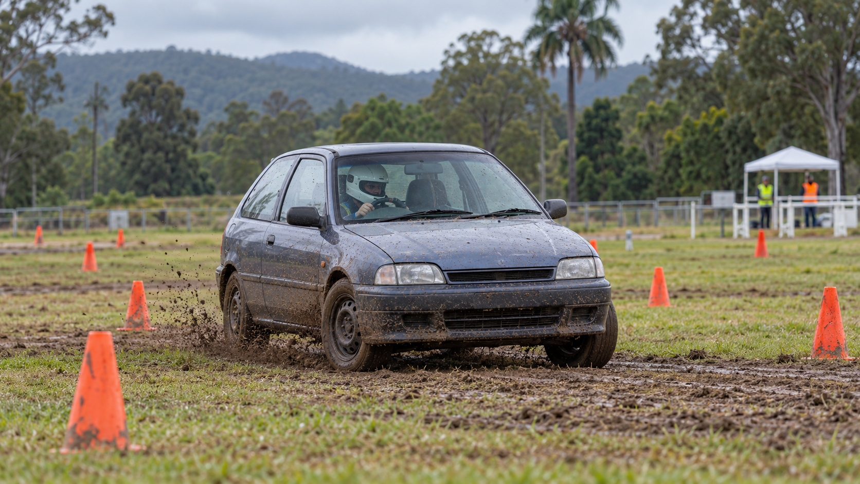 A compact competition car driving between orange cones on a wet grass khanacross course.